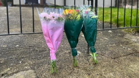 Three bunches of flowers laid at a gate in front of a flat in Bristol. One has pink packaging and the others have green packaging.