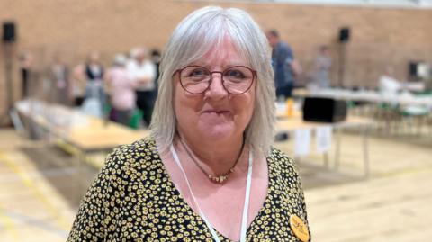 Lorne Dupre wearing a black and yellow floral dress and standing in a sport hall where an election count is taking place