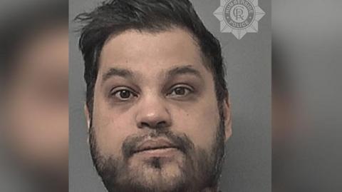 Police custody photograph of Suleman Baig. He has black hair and and beard