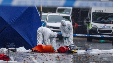Image shows forensics officers examining material in the road near to the synagogue. They are dressed in white boiler suits and are standing next to a blue tent. There are emergency vehicles in the background.