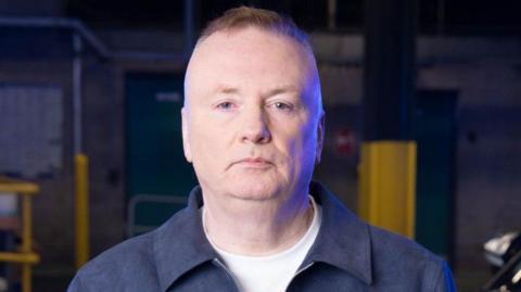 A man looks into the camera, he has short, cropped brown hair and is wearing a blue jacket and white t-shirt. He appears to be standing in a multi-storey or underground car park. 