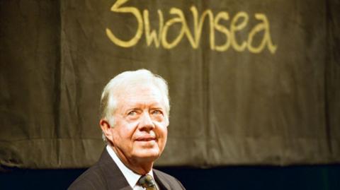 Jimmy Carter dressed in a suit with a Swansea sign behind him