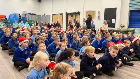 Rows of primary-age children sat on the floor of a school hall looking forward. Some are wearing santa hats. Around the room are various members of staff.