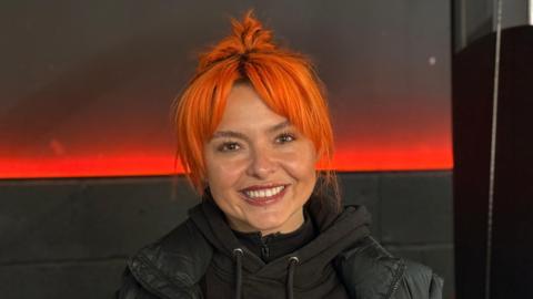 A woman, Rosie Webber, with bright orange hair smiling at the camera, wearing a black hoodie. She is inside a dimly lit gym.