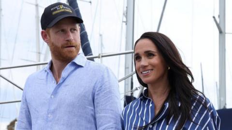 Prince Harry and Meghan walking next to each other, both in blue shirts as sails of boats are visible behind them in Sydney Harbour. Photo taken April 17 2026.