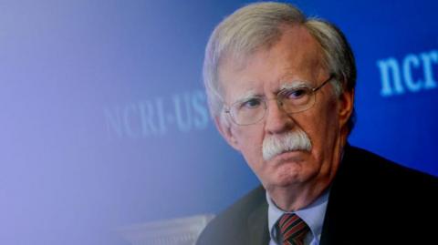 John Bolton at an event in August. He has a serious expression on his face, grey hair, a grey moustache and wire-rimmed glasses. He's wearing a black jacket, blue shirt and red and blue striped tie. There is a bright blue background.