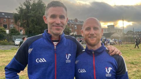 Two French GAA coaches standing on field wearing blue. 