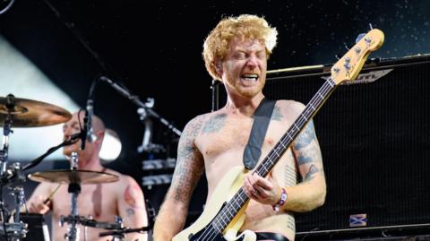James Johnston, of the band Biffo Clyro, performs on stage topless with pink trousers. His twin brother Ben, also topless, plays at a drum kit in the background.