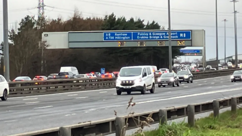 Section of M8 showing backed up traffic in the westbound lane, free flowing traffic in the eastbound lane and a sign overhead which reads: "A8 Renfrew, A736 Hillington, M8 Paisley & Glasgow Airport Erskine Bridge & Greenock"