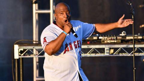 Young MC performs onstage. He is wearing a white Los Angeles jersey with red writing and is singing into a hand-held microphone