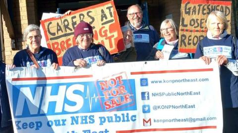Maddy Nettleship holding a banner at a protest outside the Queen Elizabeth Hospital in Gateshead. The large banner is held by three people and reads, Keep Our NHS Public. The protesters are holding banners and wearing matching T-shirts.