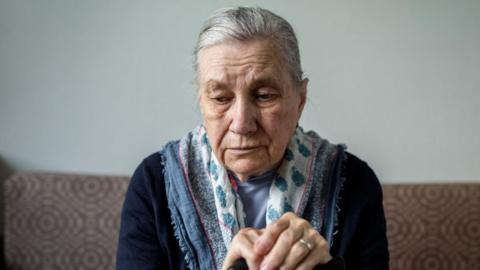 A stock image of an older woman sitting on a sofa looking down. She has longer, grey hair which has been brushed back. She is wearing a patterned scarf and has her hands placed on top of a walking cane.