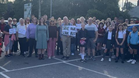 Parents, children and local politicians standing with some holding signs saying save our school bus.