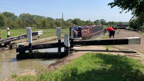 A canal lock, with a man bending over, water to the left and grass to the right. Trees are in the distance. A narrowboat is on the water, with a dog and a man at the front.