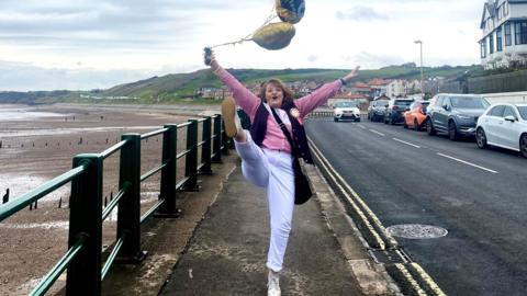 A woman holding balloons and kicking her leg up high with Sandsend beach in the background