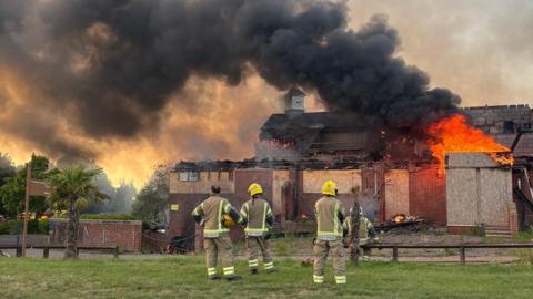 A golf club building with boarded up windows with smoke billowing upwards and part of the roof destroyed.
