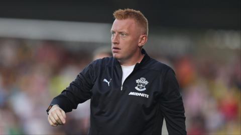 Southampton manager Will Still wears a stern focused expression on his face as he guides his players by pointing. He wears a black quarter zip top with a white-coloured Southampton FC crest.