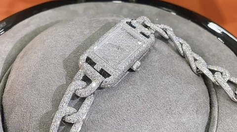 Photo of a necklace/bracelet in silvery diamond material shaped like a watch, lying on top of a grey jewellery stand.