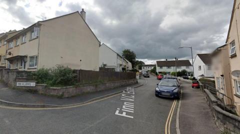 Google Street View of the Finn V.C. Estate in Bodmin with houses on both sides of the street, multiple cars parked along the road and a red mobility on the right-hand side pavement.