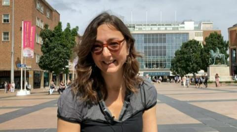 A woman is smiling at the camera, she is wearing glasses and has shoulder-length wavy brown hair. She is wearing a gret and black shirt and is sitting in Coventry city centre