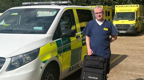 A man stood next to a van in a yellow and green ambulance livery. He is dark blue hospital scrubs and is holding a black backpack on top of a black plastic carry case.