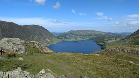A view of Crummock Water taken from the top of Rannderdale Knott, with Loweswater in the background. Crummock is surrounded by green fells. The sky is blue with a few clouds.