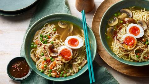 Top down view of a green bowl with mushroom ramen in. A small bowl of chilli oil sits alongside and a pair of blue chopsticks lie on the bowl.