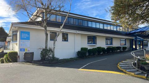 The image shows the exterior of Guernsey's Medical Specialist Group premises. It is a two-storey cream building with some small trees in front. On the wall is a blue sign which says 'Medical Specialist Group' on it. The upper level of the building has a row of large square windows along the length of the roof.