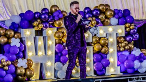 James Clarke, dressed in a purple glitter suit, on stage during Worksop's Got Talent, against a backdrop of colourful balloons and the illuminated letters WGT