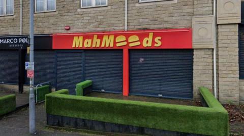 A red and yellow takeaway shopfront with the shutters down. The branding for the business reads 'Mahmoods', with two burgers instead of o's in the sign. The business colours are bright red and yellow. 