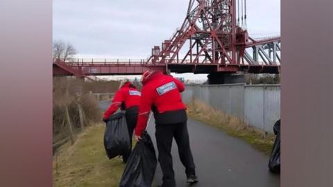 A man and a woman picking up litter and putting it into black bags while walking along a riverside path, with Middlesbrough's Newport Bridge in front of them.