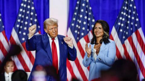 Donald Trump, wearing a blue suit and red tie, stands next to Tulsi Gabbard, in a powder blue pantsuit, on a stage full of American flags