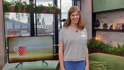 Colletta Smith, a white female, stands just right of centre wearing a grey short-sleeved top. Her blonde hair is shoulder length and she smiles at the camera. The Morning Live studio is behind her. 