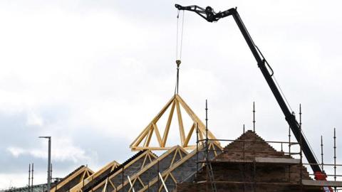 A stock image of houses being built. A crane is lowering the roof frame down onto bricks.
