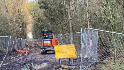 An orange digger in the middle of a muddy path at the construction site. The area is fenced off. There is a yellow sign at the start of the path.