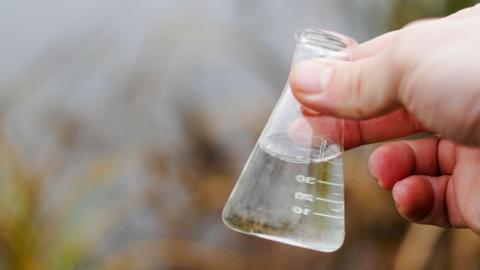 A glass beaker of water with a blurred image of a river in the background.