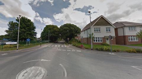 Mini-roundabout with a school to the left and a housing estate to the right/ On the road is a zebra crossing with 20mph speed limit signs. 