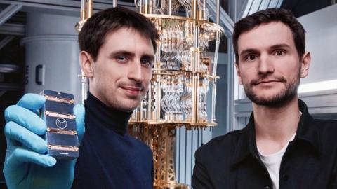 Two men in the foreground staring at the camera, one of whom is holding a black and gold device, with a large gold device visible in the background