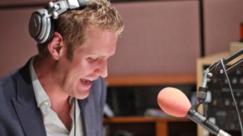 Jonathan sits behind a microphone in the studio, with a large orange foam mic cover. He is chuckling and wears headphones, with one slightly off his right ear. He wears an open necked grey shirt and blue-grey suit jacket.
