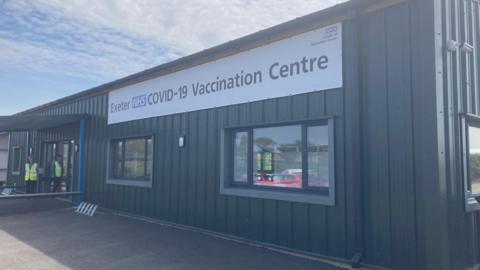 A dark green building with a large white banner which says Exeter NHS Covid-19 Vaccination Centre. Two people in high-vis vests are stood at the entrance. 