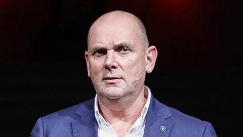 Jim Gamble. He is bald with brown eyes, wearing a blue blazer and white shirt with pin on his lapel. He is wearing a small earpiece microphone and the background is black.