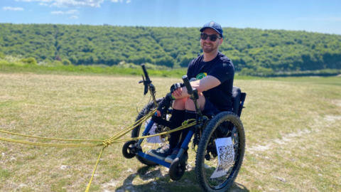 Dale Muffett is in a wheelchair smiling at the camera. He is wearing shorts and a t-shirt, has a navy cap on and dark sunglasses.