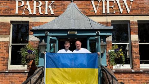 Two men are holding a Ukrainian flag and standing outside a hotel.