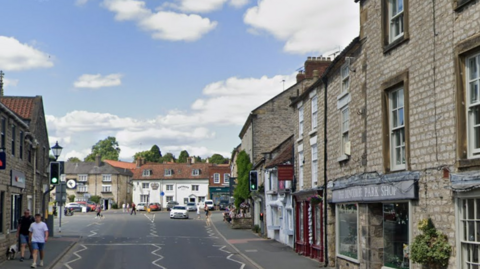 Bridge Street, Helmsley as it approaches the Market Place. A road in a market town with a range of pale stone buildings on either side