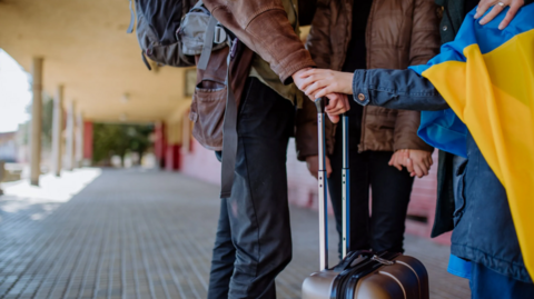 Lower body images of two people wearing brown jackets, greeting someone wearing a Ukraine flag around their shoulders. They appear to be standing at a station.