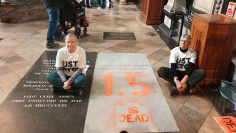 Two Just Stop Oil are seen sitting on the floor in Westminster Abbey at each side of Darwin's marble nave wearing white t-shirts with the group's name on it. "1.5 is dead" is painted on in large orange font.