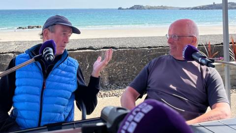 Two men in front of microphones discussing politics, the man on the left is wearing a blue gillet and is gesticulating towards the man on the right. They are in front of a beach backdrop on a sunny day. 