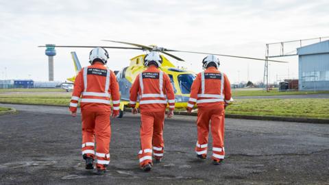 Three figures in orange uniforms and white helmets walk towards a yellow helicopter at an airport.