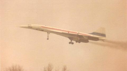 The Concorde prototype is seen taking off into the air. It is white with a red and a blue stripe.