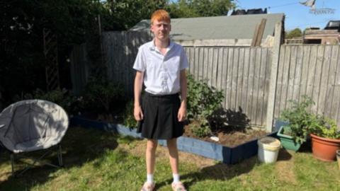 Young boy wearing white polo top and school style grey skirt standing in a back garden with grass and plant pots